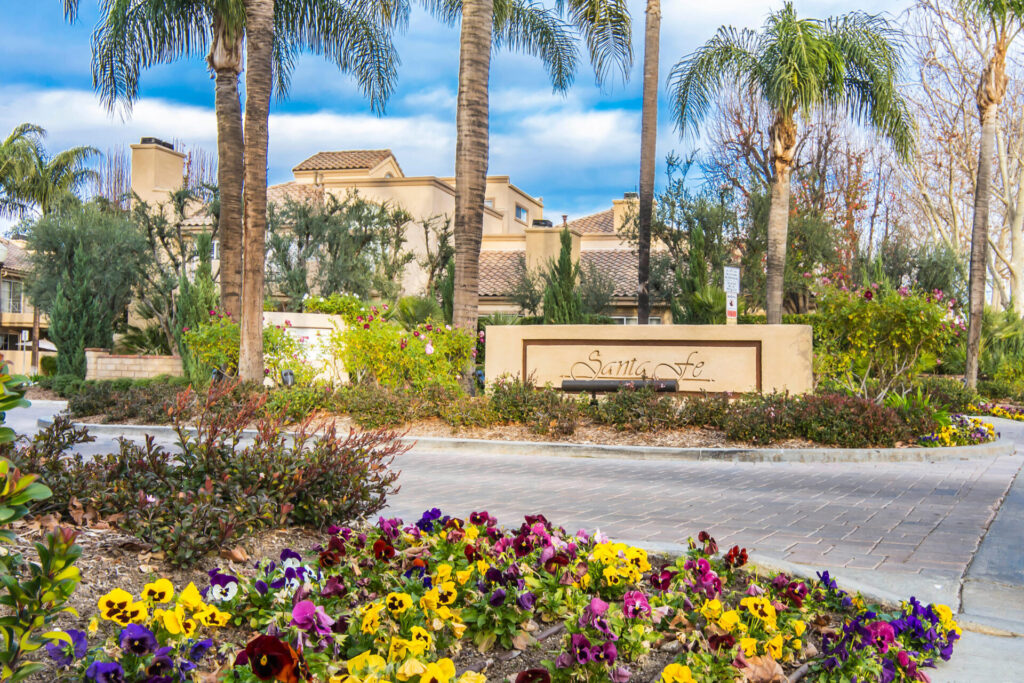 The entrance to the Santa Fe Condominium Complex in Valencia, California, showing a brick entrance with lush flowers and tall palm trees.